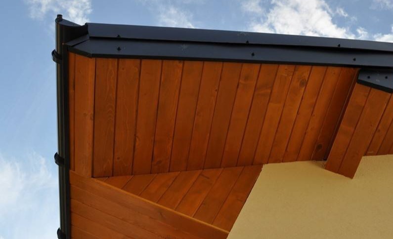 Drewniana podbitka dachowa w kolorze miodowego brązu z czarnymi elementami orynnowania na tle błękitnego nieba z chmurami.