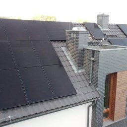 SUPER OZE SPÓŁKA Z OGRANICZONĄ ODPOWIEDZIALNOŚCIĄ - Czarny panel fotowoltaiczny na szarym, spadzistym dachu domu jednorodzinnego z ceglanym kominem i oknem dachowym, widoczne inne domy w tle.
