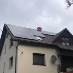 Dom jednorodzinny z zamontowanymi panelami słonecznymi na dachu, widoczne okno dachowe i komin z cegły, pochmurne niebo w tle.