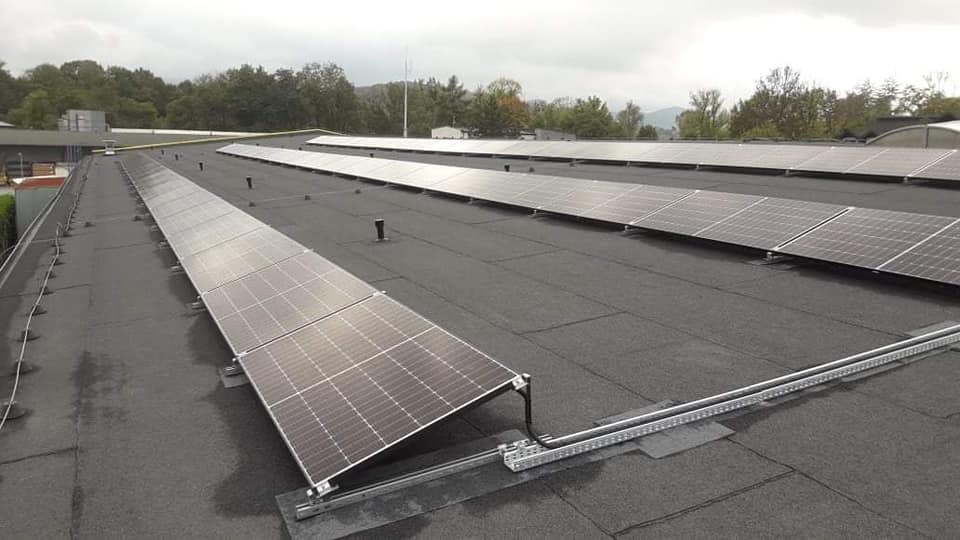 Rząd paneli słonecznych zamontowanych na płaskim, bitumicznym dachu budynku, widoczne mocowania i szyny montażowe, w tle zieleń drzew i pochmurne niebo.