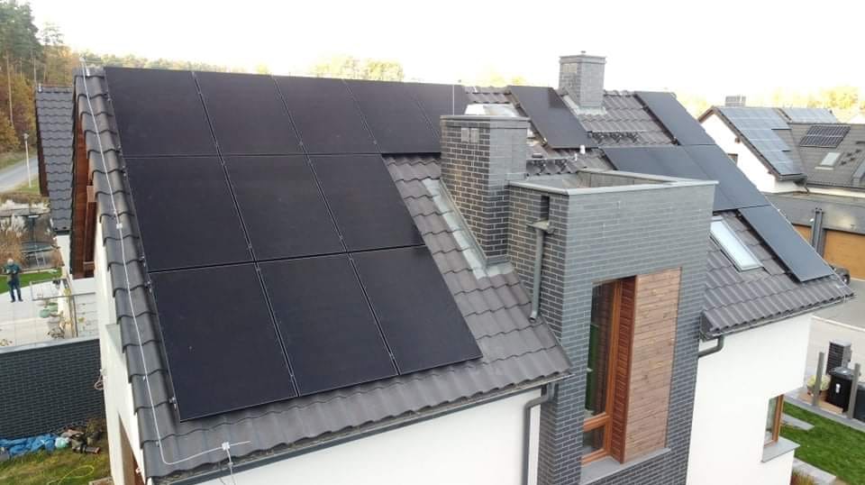 Czarny panel fotowoltaiczny na szarym, spadzistym dachu domu jednorodzinnego z ceglanym kominem i oknem dachowym, widoczne inne domy w tle.