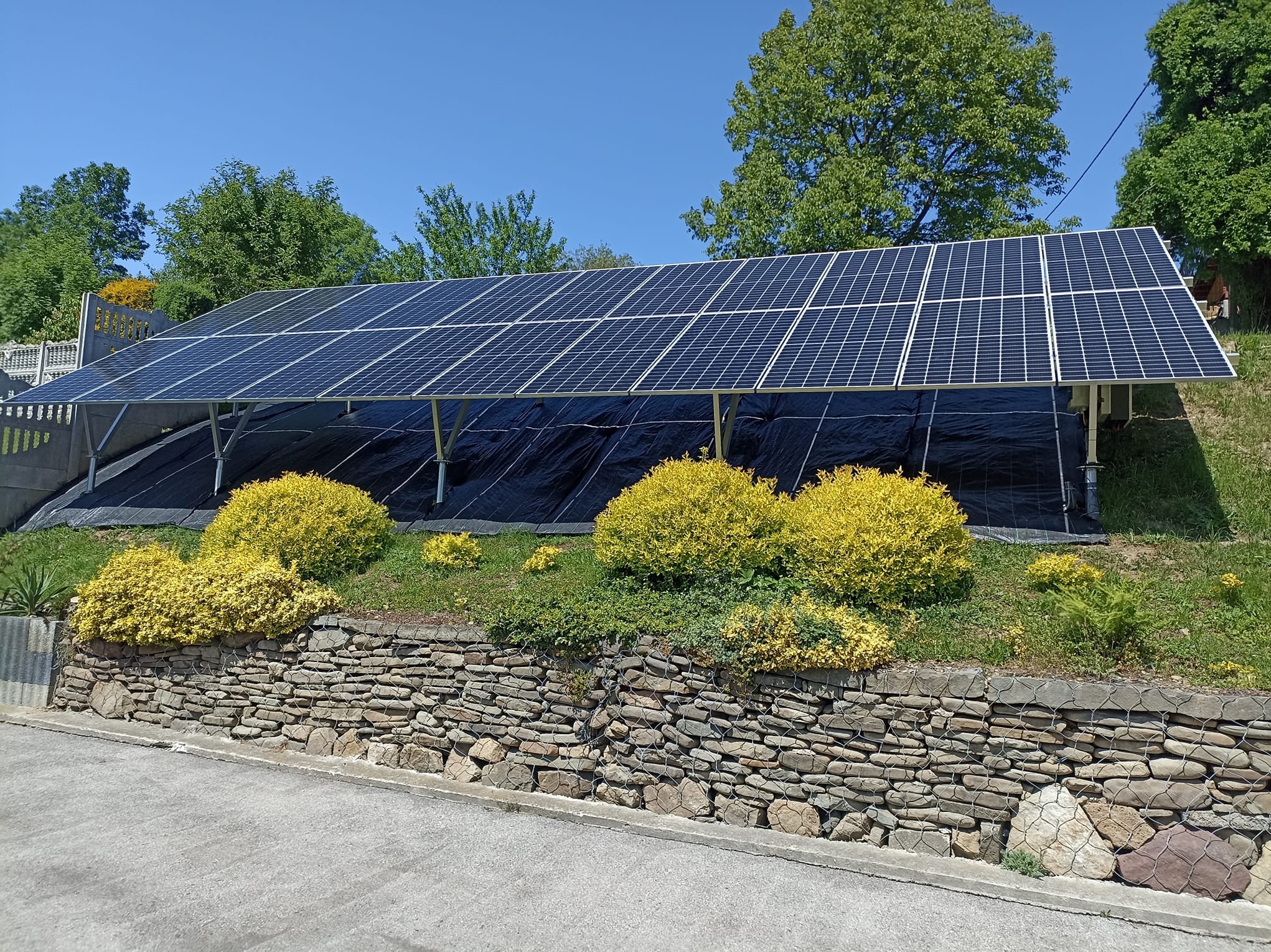 Instalacja fotowoltaiczna na stelażu, zabezpieczona czarną folią od spodu, widoczna z dołu, z ozdobnymi krzewami i kamiennym murkiem na pierwszym planie, błękitne niebo w tle.