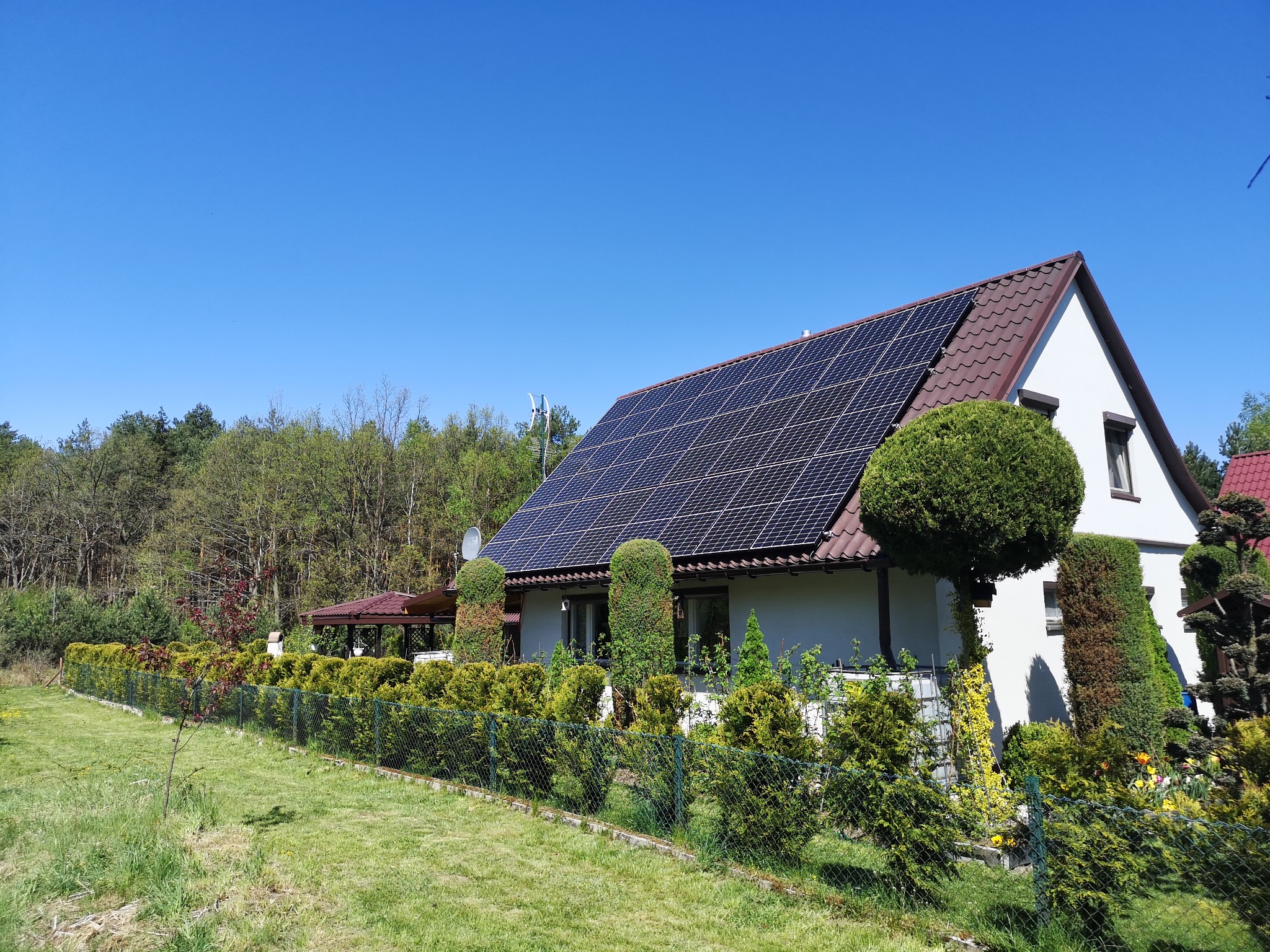 Dom jednorodzinny z brązowym dachem pokrytym panelami słonecznymi, otoczony zielonym ogrodem z ozdobnymi krzewami i metalową siatką ogrodzeniową w słoneczny dzień.