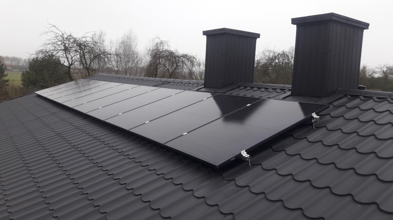 Ciemne panele słoneczne zamontowane na ciemnym, metalowym dachu z dwoma kominami w tle pochmurnego nieba i drzew bez liści.