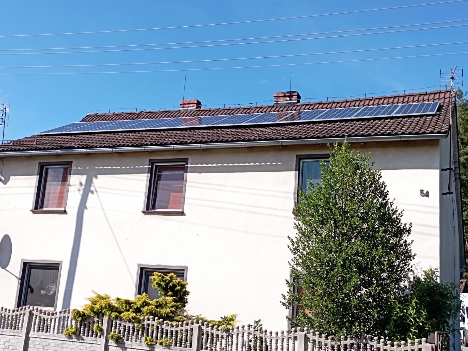 Biały dom jednorodzinny z brązowym dachem, na którym zamontowano panele słoneczne; widoczne kominy, instalacja odgromowa, antena telewizyjna oraz częściowo przysłaniające fasadę drzewo i krzewy.