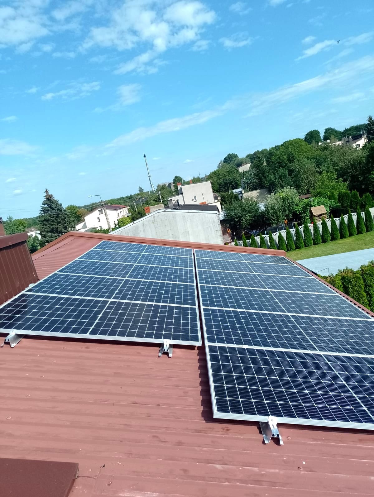 Panele fotowoltaiczne zamontowane na metalowym dachu w kolorze ceglastym, widok z góry, w tle zabudowa jednorodzinna i błękitne niebo z chmurami.