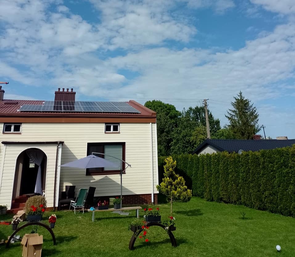 Dom jednorodzinny z panelami słonecznymi na dachu, widok z ogrodu z parasolem i meblami ogrodowymi, jasne niebo z chmurami.