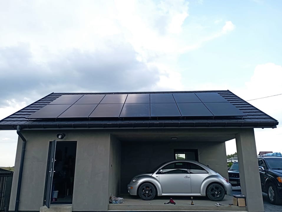 Czarny dach z panelami fotowoltaicznymi na szarym budynku z garażem, w którym stoi srebrny Volkswagen Beetle, z widocznymi narzędziami i kartonem na podłodze.