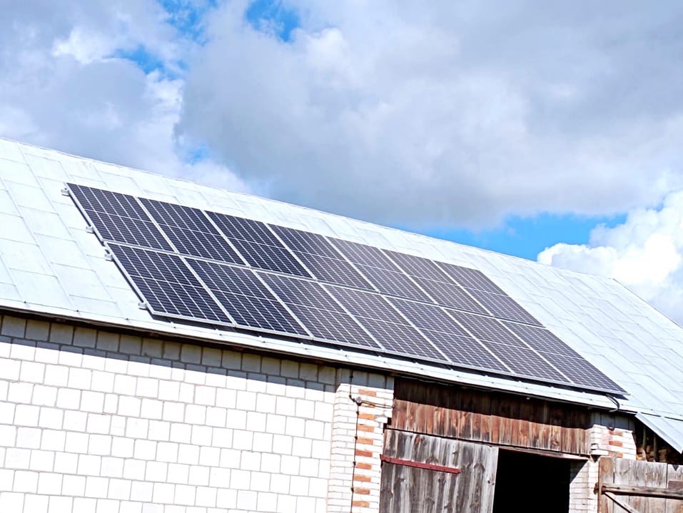 Panel słoneczny na dachu budynku gospodarczego z białej cegły i drewnianymi drzwiami, błękitne niebo z chmurami w tle.