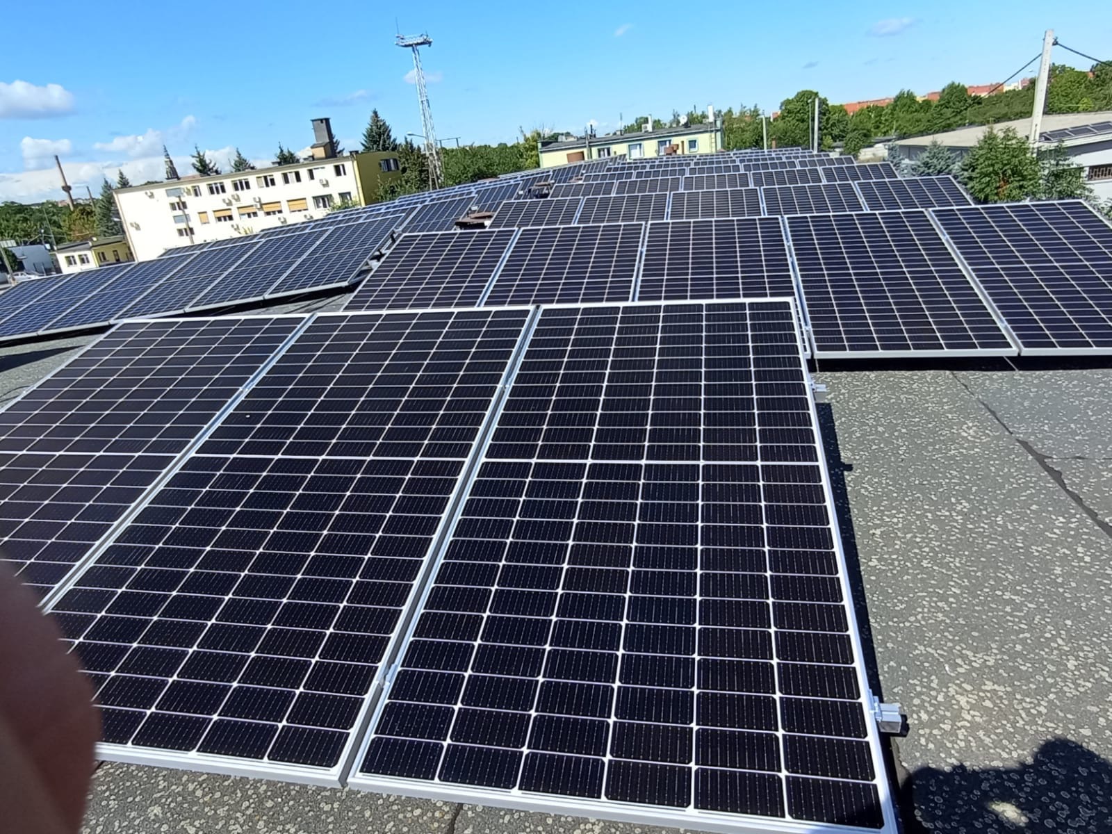 Rząd paneli fotowoltaicznych na płaskim dachu budynku, widoczne budynki w tle i błękitne niebo z lekkimi chmurami.