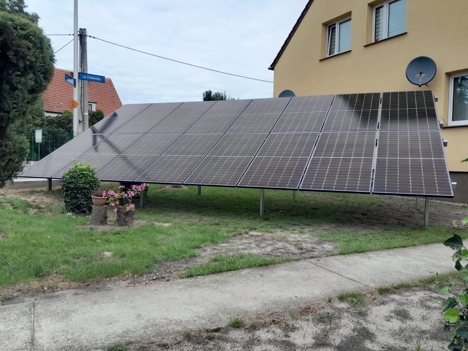 Panel słoneczny na metalowych podporach ustawiony w ogrodzie przed domem z żółtą elewacją, widoczny znak uliczny 'ul. Pawłowicka'.