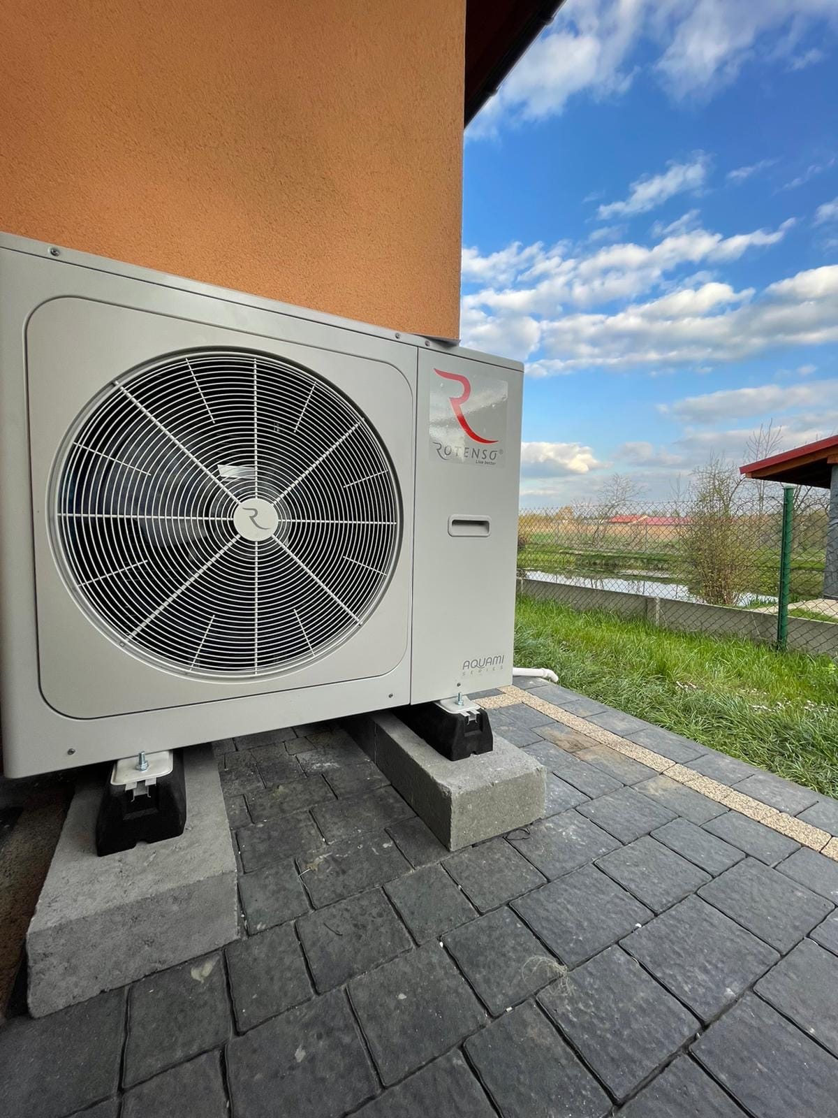 Biała pompa ciepła marki Rotenso Aquami, zamontowana na betonowych podstawach obok budynku z pomarańczową elewacją, na tle błękitnego nieba z chmurami.
