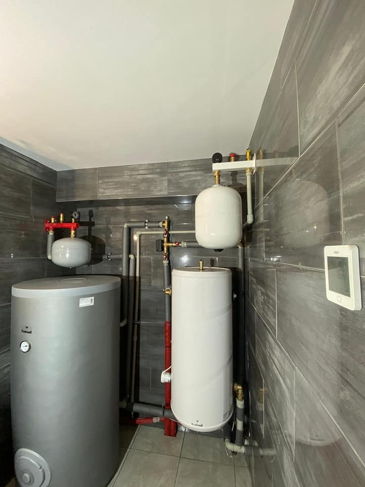 Zestaw dwóch zbiorników buforowych, szary i biały, z widoczną instalacją hydrauliczną w pomieszczeniu wyłożonym szarymi płytkami imitującymi kamień.