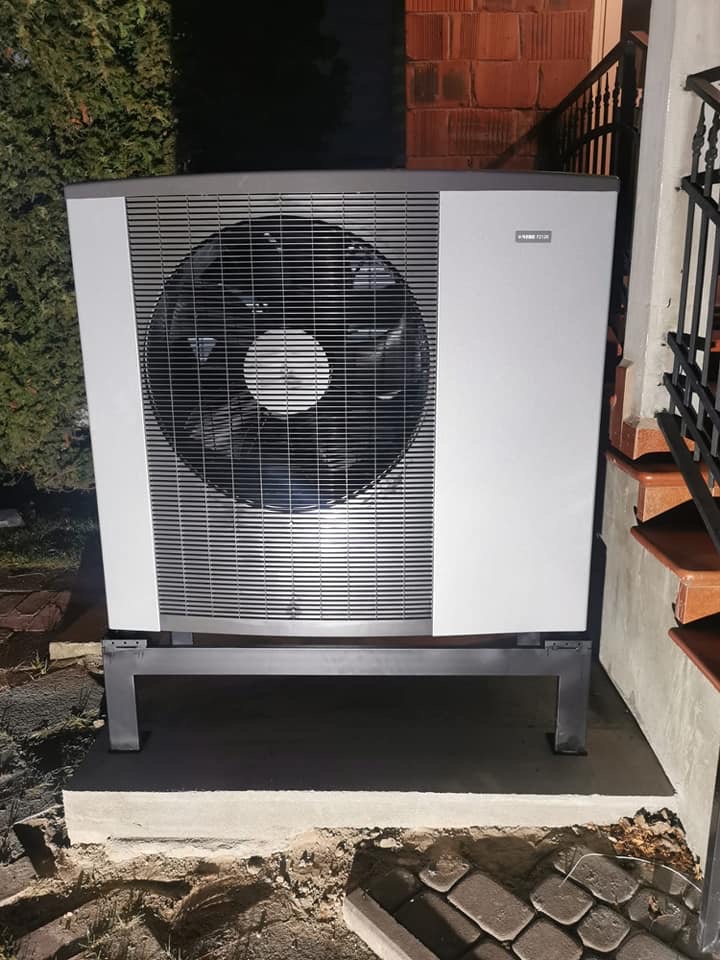 Zewnętrzna jednostka pompy ciepła powietrze-woda, zamontowana na betonowym fundamencie obok schodów z ceglanymi stopniami i metalową barierką, otoczona zielenią i kostką brukową.