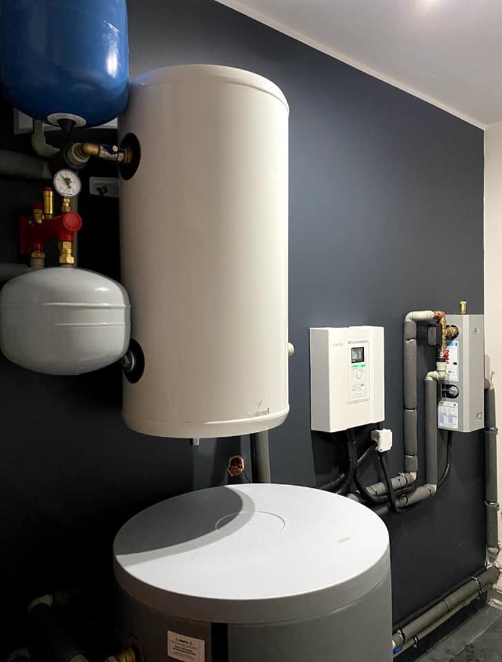 Kompletna instalacja grzewcza z zbiornikami akumulacyjnymi, armaturą i sterowaniem elektronicznym na tle ciemnej ściany.