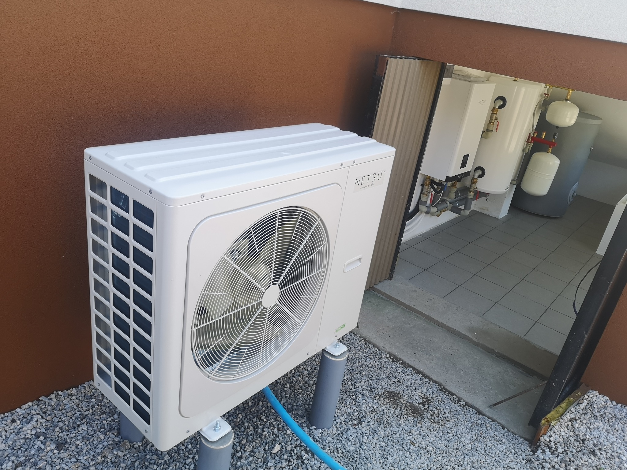 Biała jednostka zewnętrzna z wentylatorem, zamontowana na szarych wspornikach na żwirowym podłożu, z widocznym niebieskim przewodem, obok brązowej ściany budynku. W tle, przez otwarte drzwi...