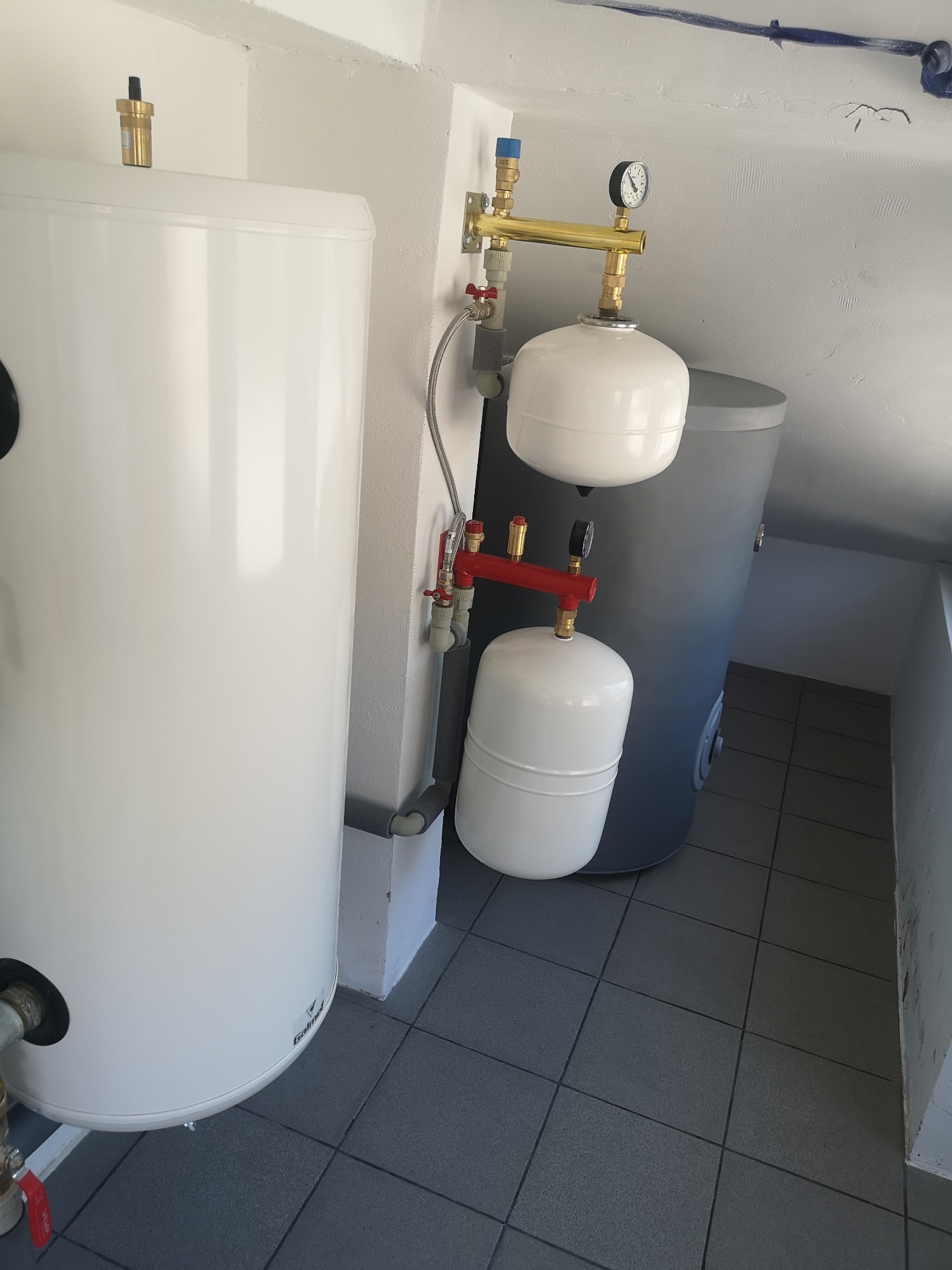 Biały zasobnik ciepłej wody użytkowej marki Galmet z osprzętem, widoczne manometry, zawory i zbiorniki wyrównawcze, szare płytki podłogowe.