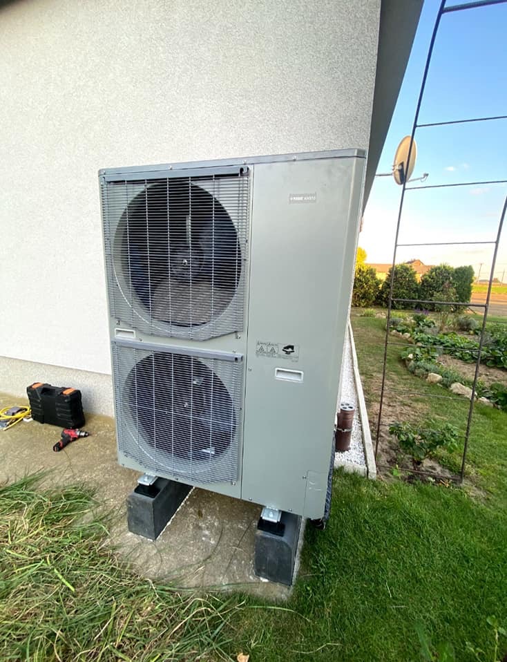 Zewnętrzna jednostka pompy ciepła, dwuwentylatorowa, zamontowana na betonowej podstawie w ogrodzie, widoczne narzędzia i elementy instalacyjne obok.