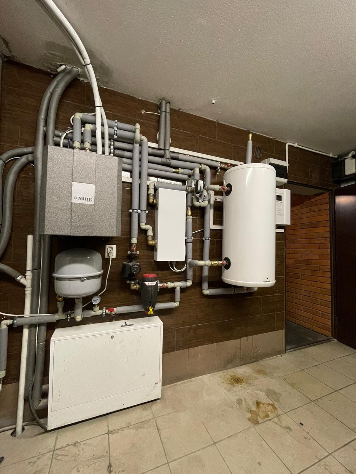 Instalacja grzewcza w kotłowni z widocznym zasobnikiem ciepłej wody, rozdzielaczem, zbiornikiem wyrównawczym oraz rurami w izolacji, na brązowej ścianie z płytek, z logo NIBE na jednym z urządzeń.