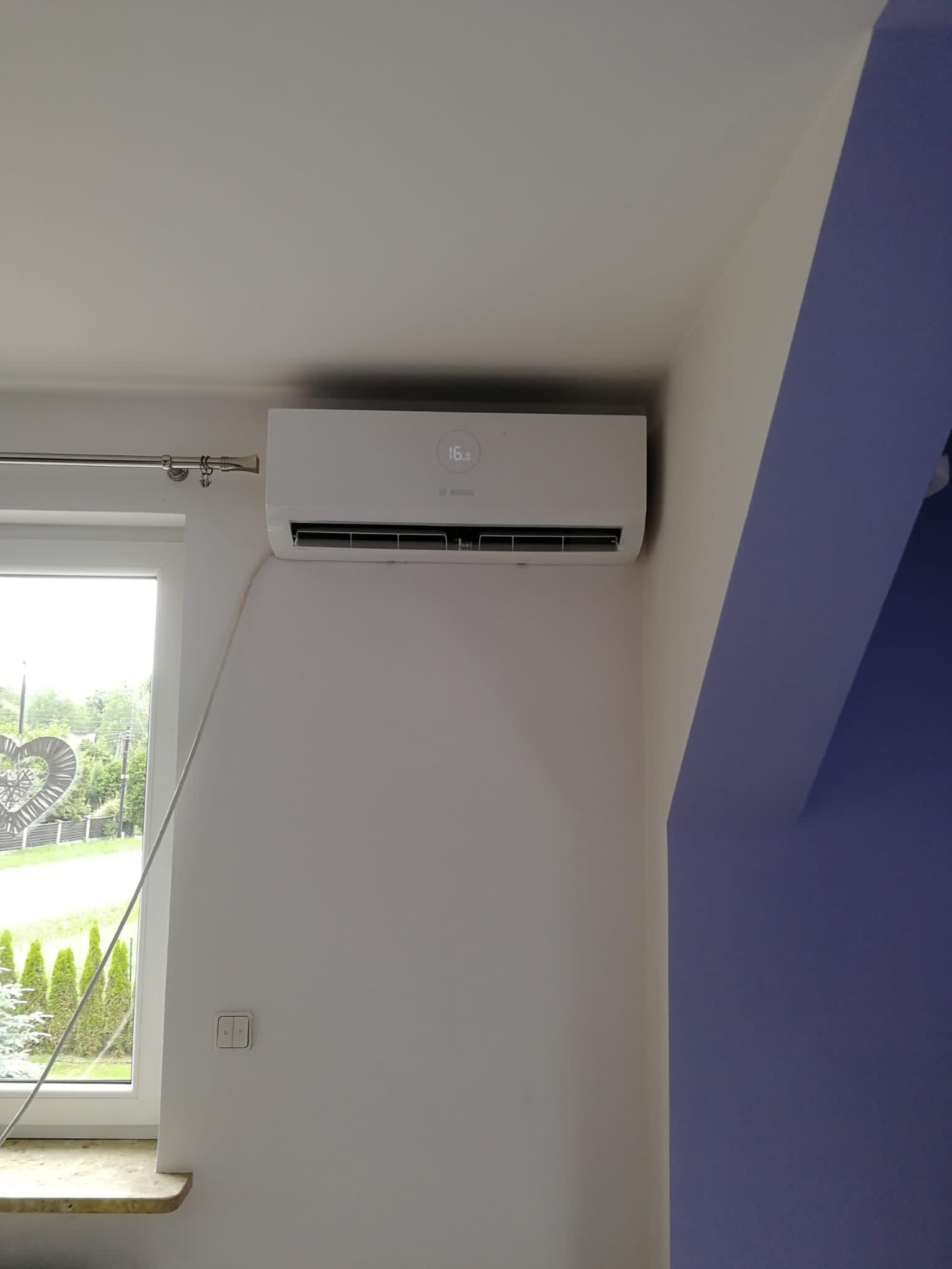 Biała klimatyzacja Bosch zamontowana na ścianie w pokoju z oknem i fragmentem ściany pomalowanej na fioletowo. Na wyświetlaczu klimatyzacji widoczna temperatura 16 stopni Celsjusza.