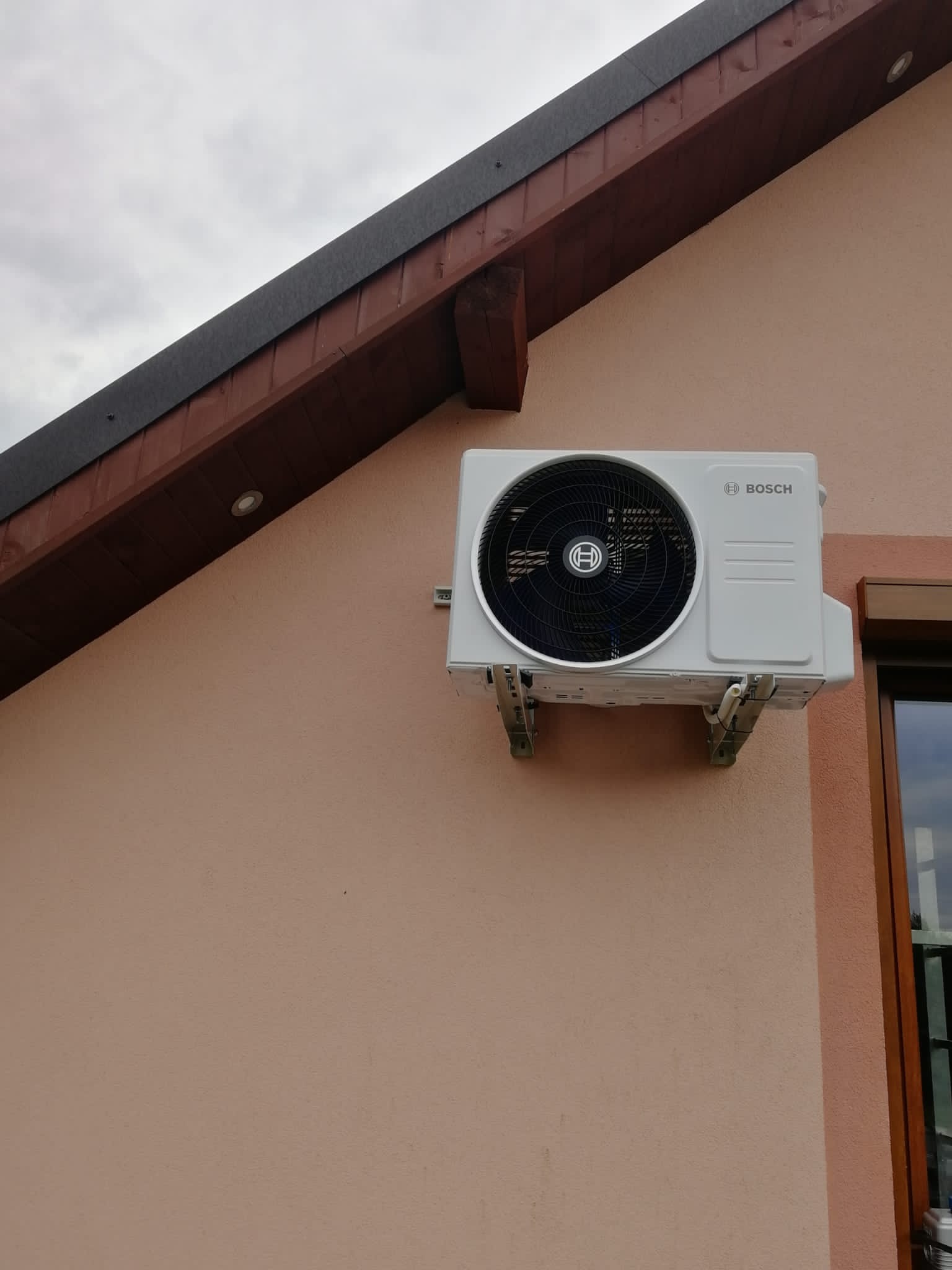 Biała jednostka zewnętrzna klimatyzacji marki Bosch zamontowana na elewacji budynku z beżowym tynkiem, widoczna część okna z brązową ramą i fragment dachu z drewnianą konstrukcją.