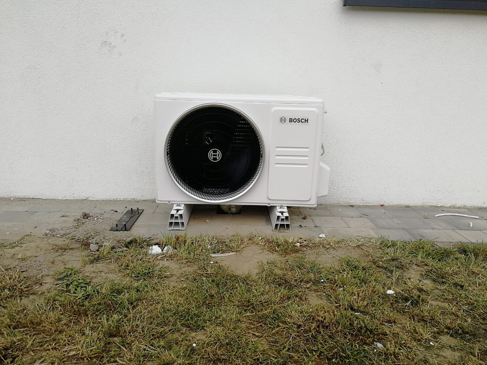 Biała zewnętrzna jednostka klimatyzacyjna Bosch na aluminiowych wspornikach przy ścianie budynku, widoczne fragmenty trawnika i chodnika z kostki.