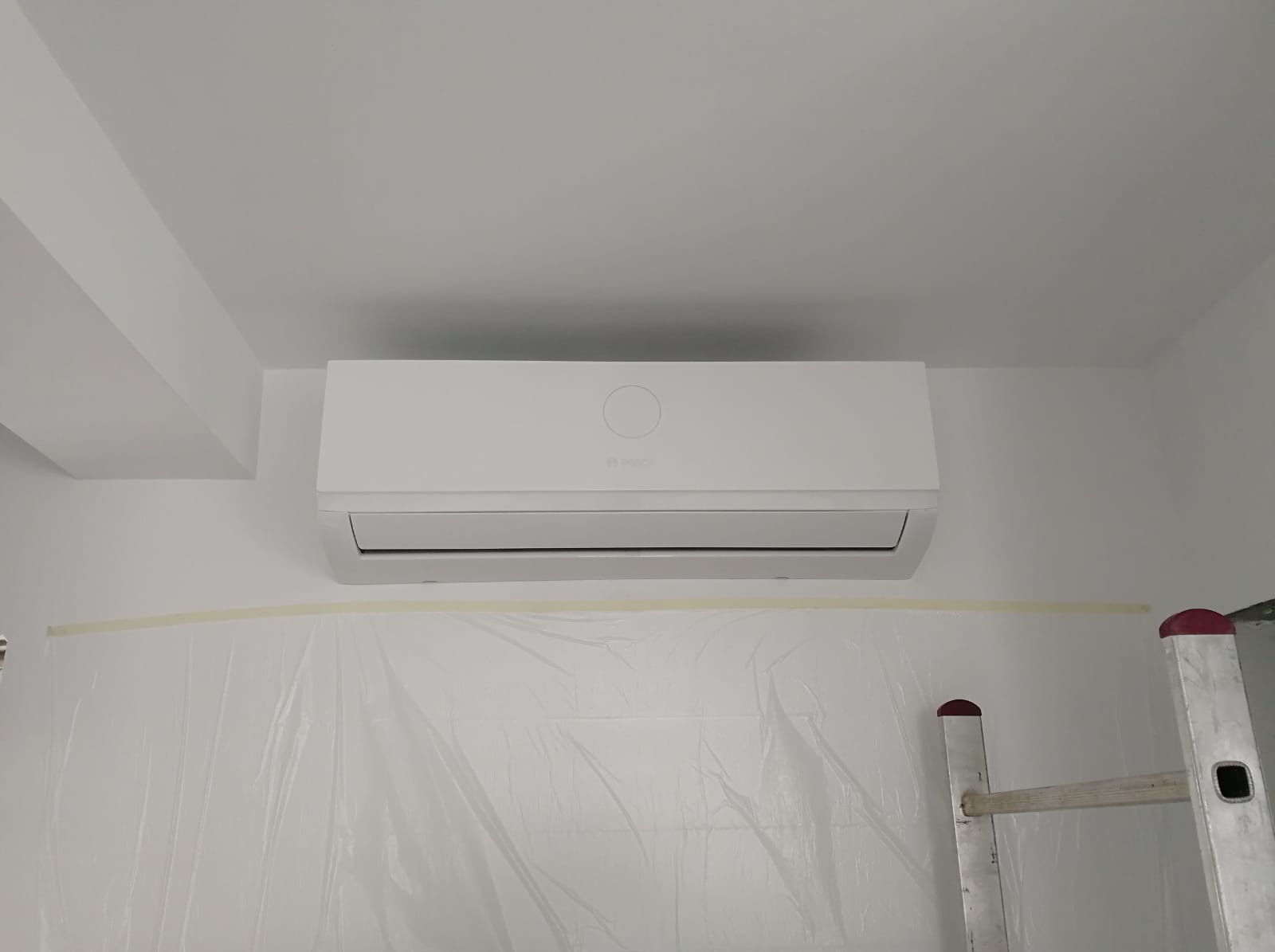 Biały klimatyzator marki Bosch zamontowany na ścianie, zabezpieczonej folią malarską z żółtą taśmą, widoczny fragment drabiny malarskiej.