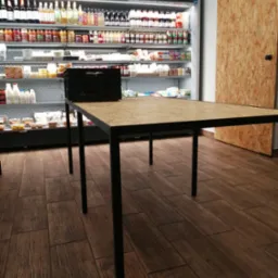Stół z blatem z płyty OSB i czarnymi metalowymi nogami, ustawiony w sklepie spożywczym z pełnymi półkami i przesuwnymi drzwiami z płyty OSB w tle.
