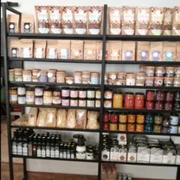 Metalowy regał z półkami wypełnionymi różnorodnymi produktami spożywczymi: kawą w ziarnach, granolą, miodami w słoikach, makaronem, olejami oraz kaszami i płatkami, ustawionymi w sklepie...