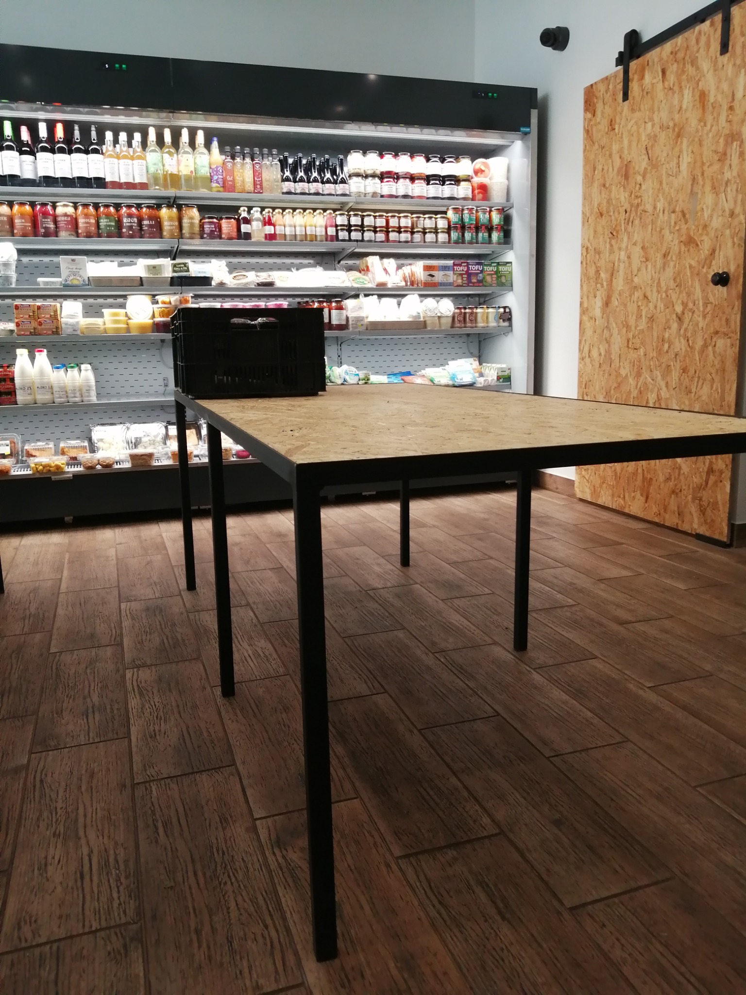 Stół z blatem z płyty OSB i czarnymi metalowymi nogami, ustawiony w sklepie spożywczym z pełnymi półkami i przesuwnymi drzwiami z płyty OSB w tle.