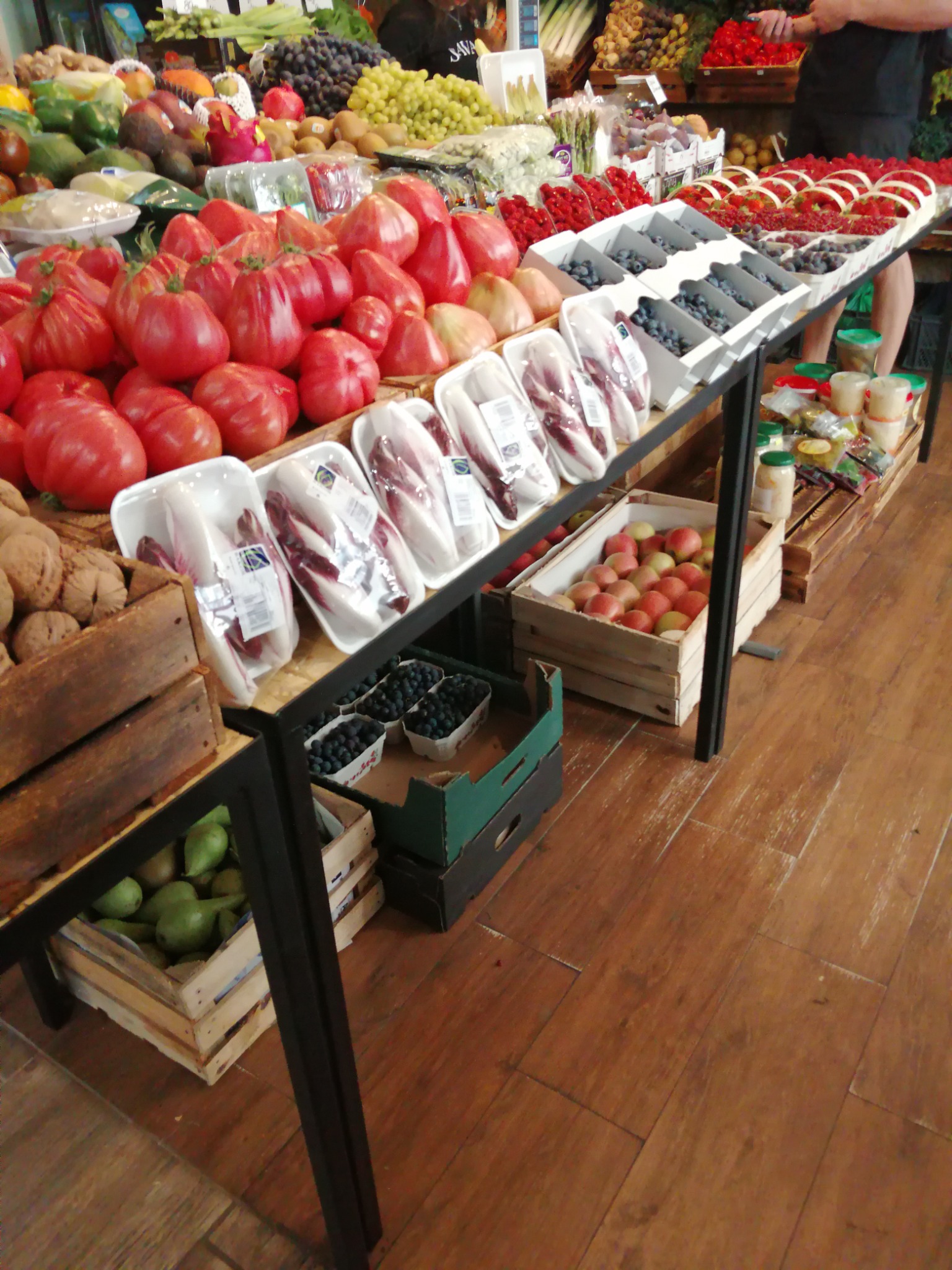 Stoisko na targu z różnorodnymi owocami i warzywami, w tym pomidory, sałata radicchio, borówki i jabłka, prezentowane w drewnianych skrzynkach i białych pojemnikach.