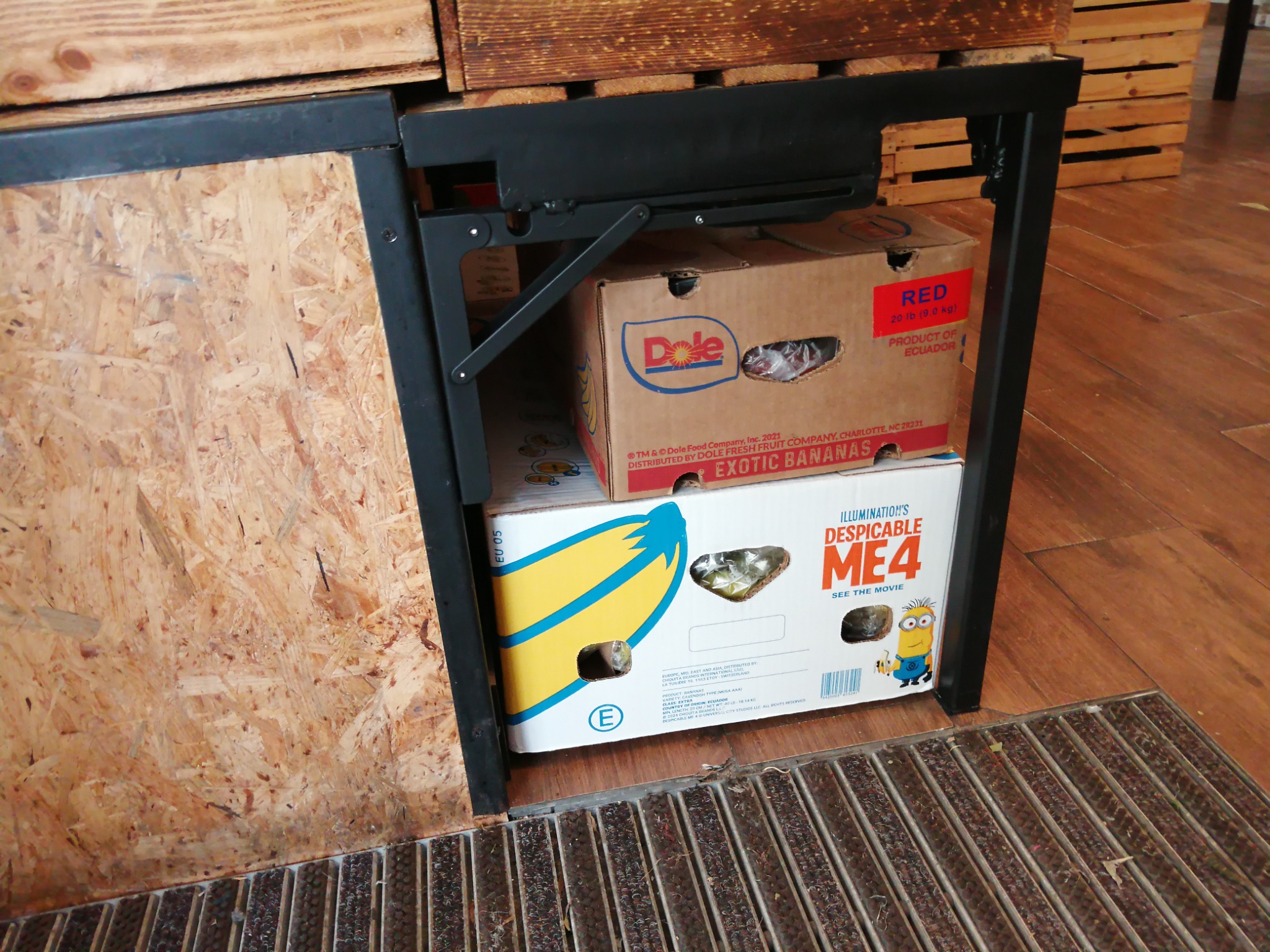 Metalowa konstrukcja z czarnych profili, prawdopodobnie fragment stołu warsztatowego lub regału, z widocznym fragmentem drewnianej ściany z płyty OSB i kratką podłogową, pod spodem ustawione...