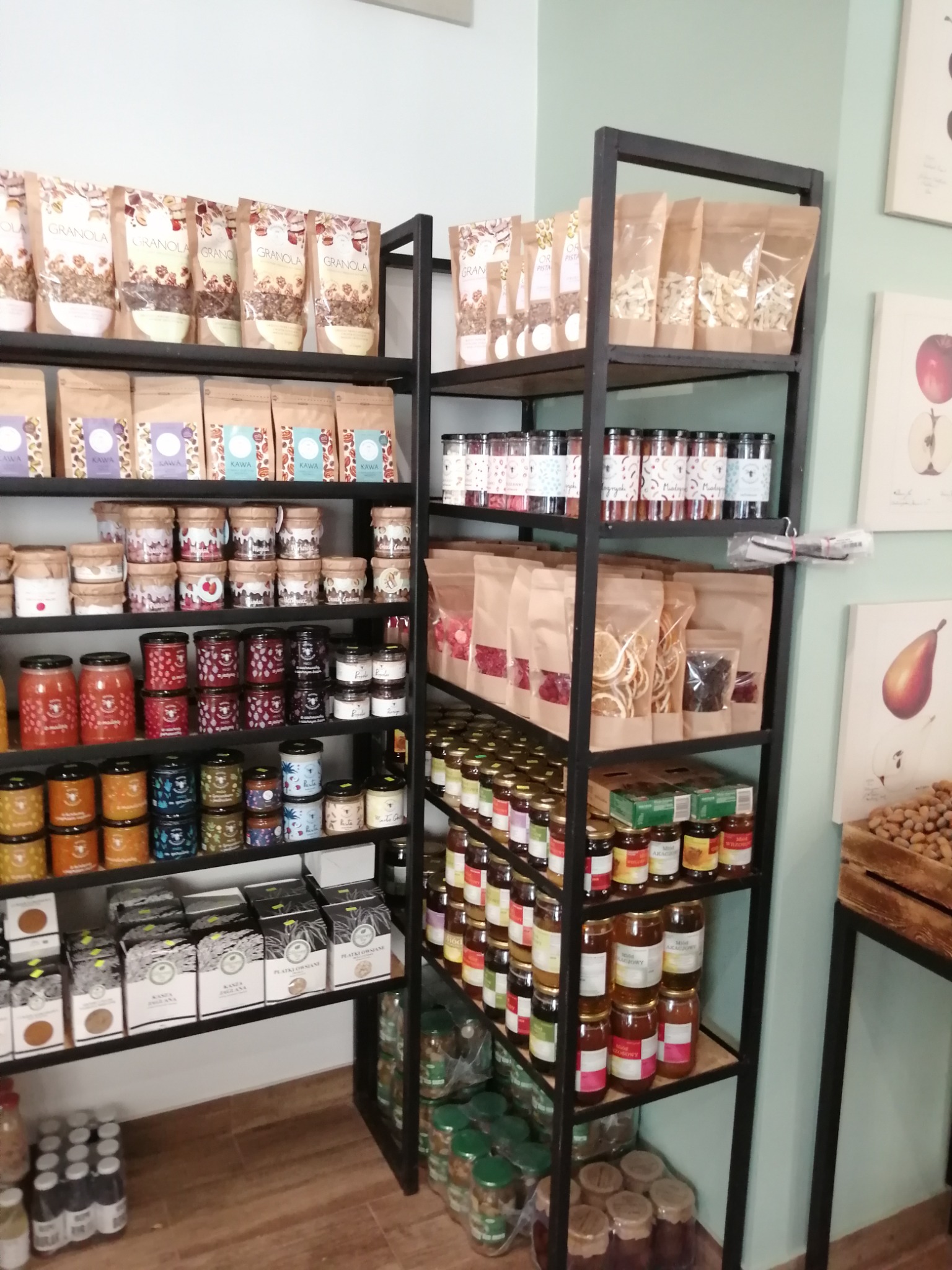 Półki regału w sklepie spożywczym wypełnione różnorodnymi produktami w słoikach, torebkach i pudełkach, w tym miodami, dżemami, granolą i płatkami owsianymi.