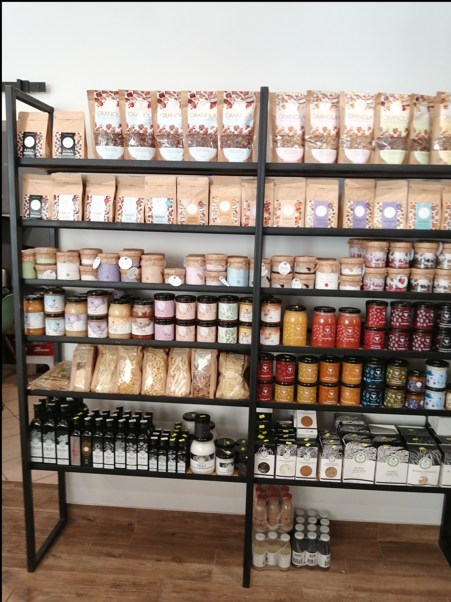 Metalowy regał z półkami wypełnionymi różnorodnymi produktami spożywczymi: kawą w ziarnach, granolą, miodami w słoikach, makaronem, olejami oraz kaszami i płatkami, ustawionymi w sklepie...