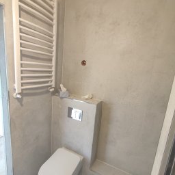 Palina Matys Matbuiding - Nowoczesna łazienka w trakcie remontu: podwieszana toaleta z szarymi płytkami imitującymi beton, biały grzejnik drabinkowy, widoczny otwór instalacyjny w ścianie.