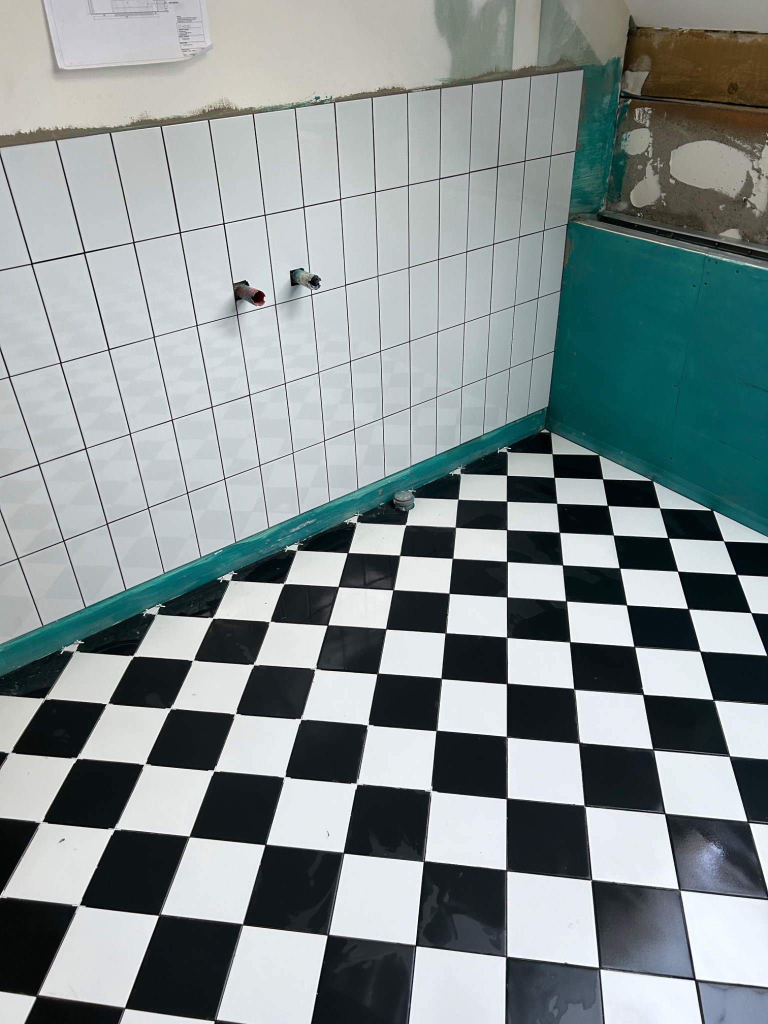 Fragment łazienki w trakcie prac wykończeniowych: białe, prostokątne płytki ścienne z czarną fugą, czarno-biała szachownica na podłodze, widoczne fragmenty instalacji wodnej i zielona hydroizolacja.