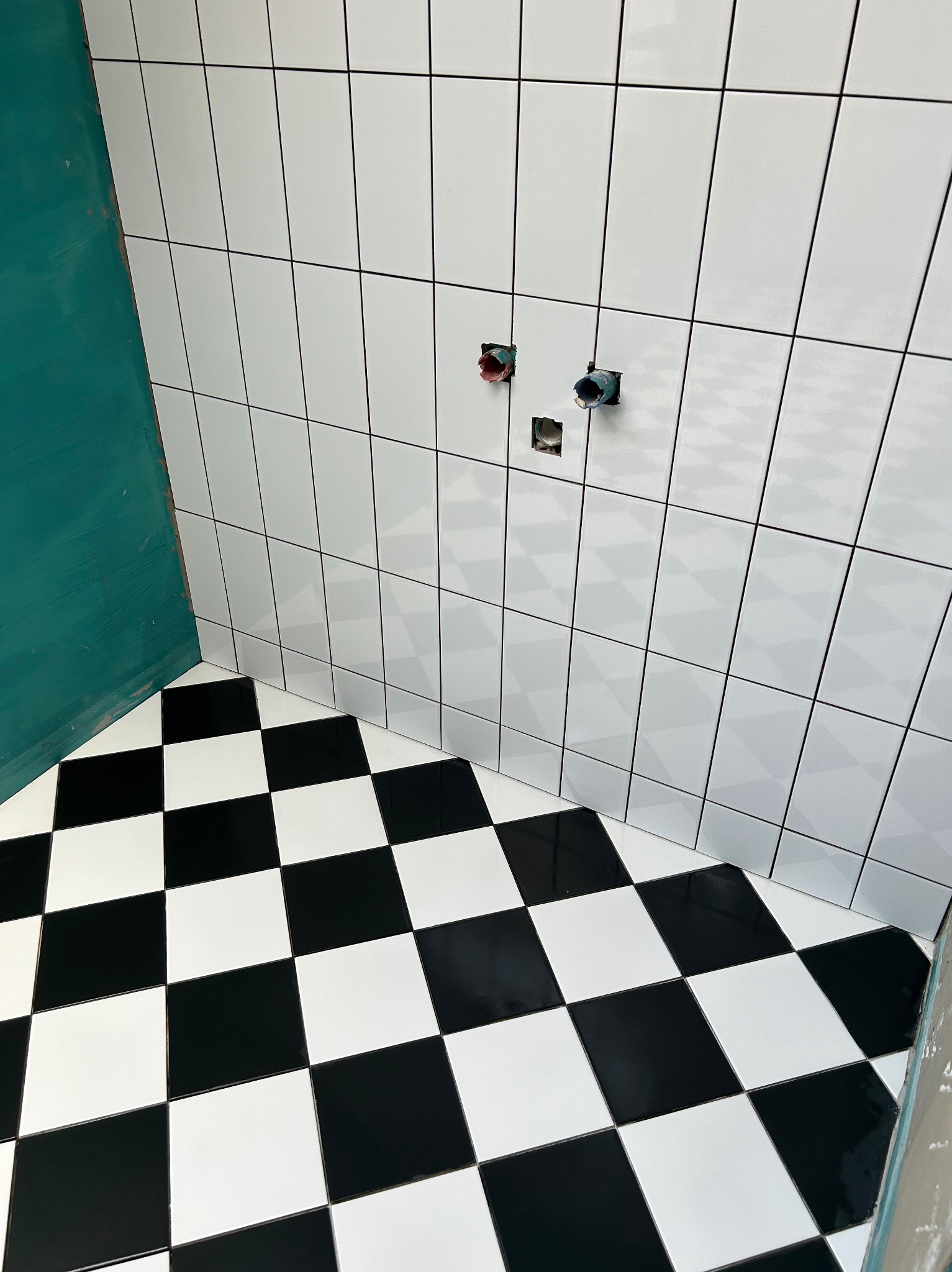Świeżo wykafelkowana łazienka z biało-czarną szachownicą na podłodze, ściany wyłożone białymi, prostokątnymi płytkami, widoczne otwory na armaturę i fragment pomalowanej na zielono ściany.