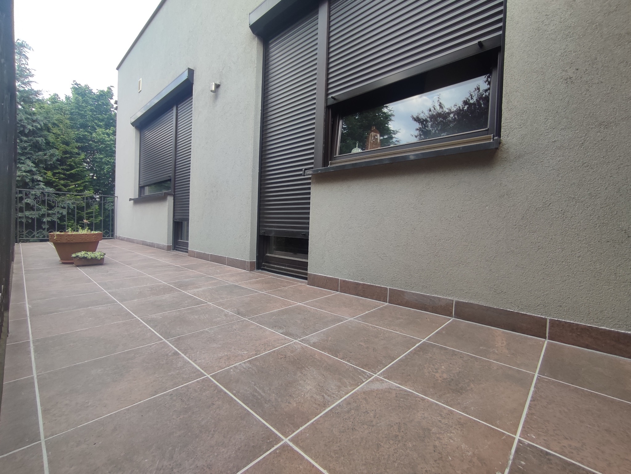 Zewnętrzny taras wyłożony brązowymi płytkami, z widocznymi roletami okiennymi i roślinami w donicach, zieleń w tle.