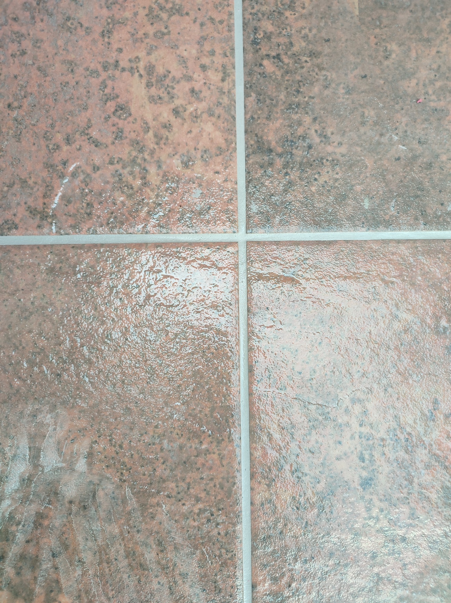 Błyszcząca, pomarańczowo-brązowa płytka podłogowa z ciemnymi plamkami i jasnoszarą fugą, ułożona w kwadraty.