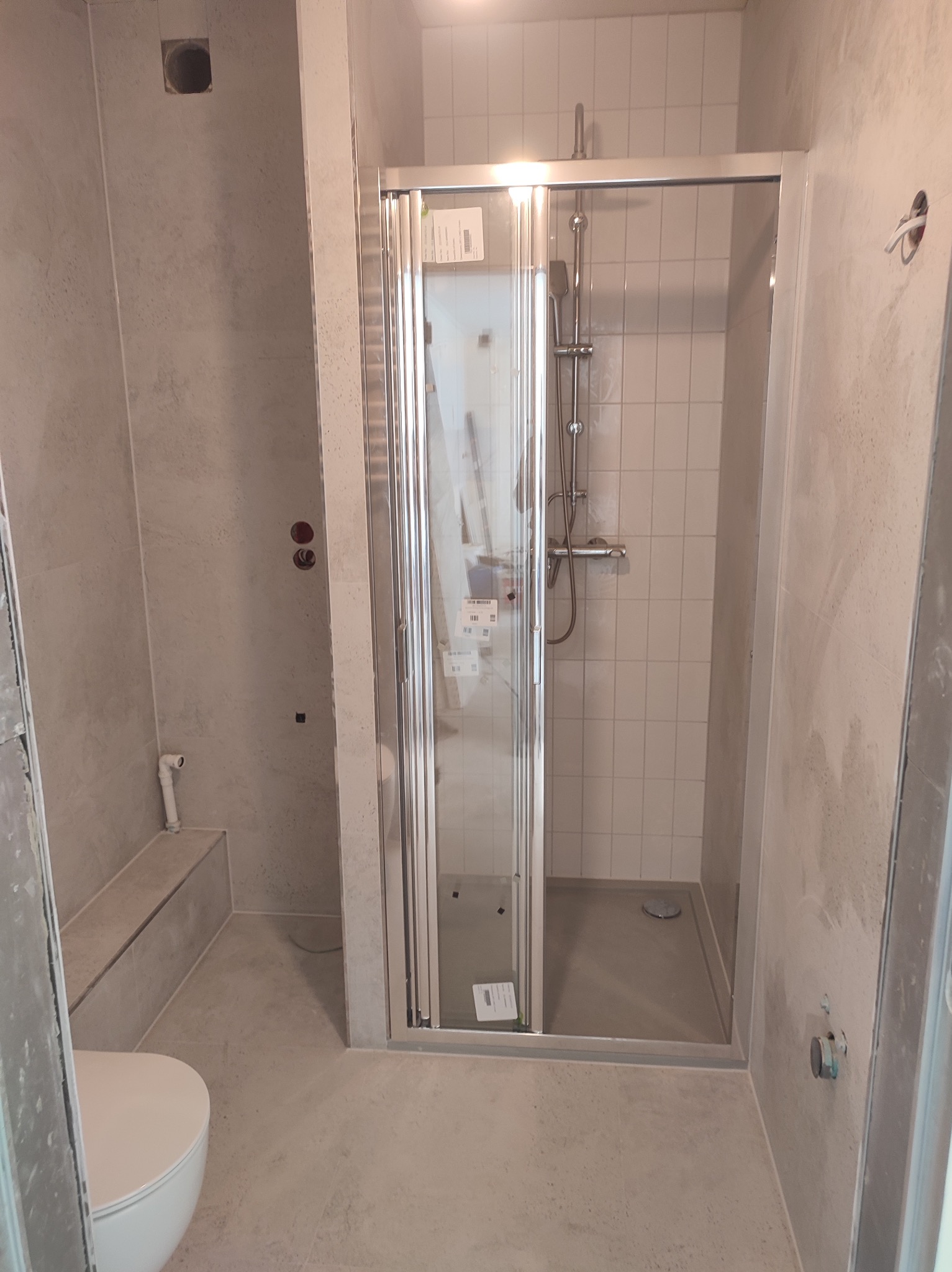 Wnętrze łazienki w trakcie wykańczania, widoczna kabina prysznicowa z przesuwnymi drzwiami, częściowo zamontowana toaleta oraz ściany pokryte szarym tynkiem strukturalnym, z widocznymi otworami...