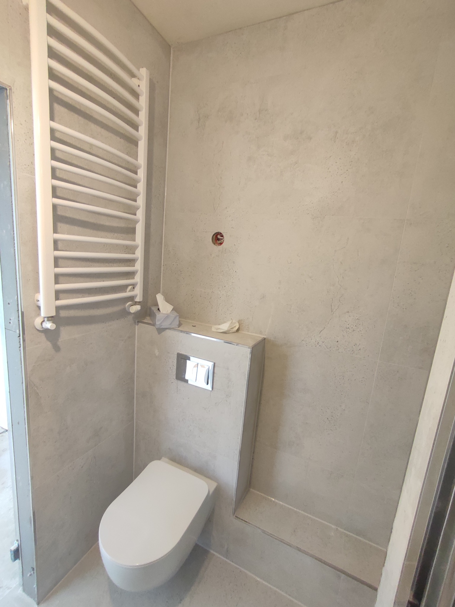 Nowoczesna łazienka w trakcie remontu: podwieszana toaleta z szarymi płytkami imitującymi beton, biały grzejnik drabinkowy, widoczny otwór instalacyjny w ścianie.