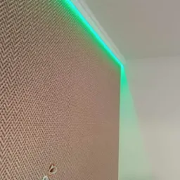 Beżowa tapeta w jodełkę z podświetleniem LED RGB wzdłuż sufitu, widoczne gniazdko elektryczne na ścianie.