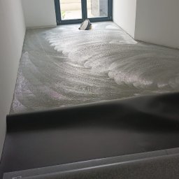 Marek Turowski Usługi Ogólnobudowlane - Przygotowanie podłoża pod nową szarą wykładzinę podłogową, widoczne resztki kleju i narzędzia w pustym pomieszczeniu z dużym oknem balkonowym.