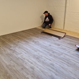 Marek Turowski Usługi Ogólnobudowlane - Pracownik w czarnej bluzie i czapce układa panele podłogowe w jasnym pomieszczeniu. Część podłogi jest już pokryta panelami o wzorze drewna, a obok leży otwarte opakowanie z panelami.