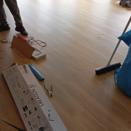 Marek Turowski Usługi Ogólnobudowlane - Montaż paneli podłogowych w jasnym pokoju z oknami balkonowymi, widoczne narzędzia, paczki paneli Gerflor Creation 55 i pracownicy podczas sprzątania po montażu.