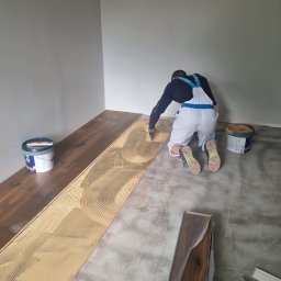 Marek Turowski Usługi Ogólnobudowlane - Parkieciarz rozprowadza klej szpachelką na podłożu pod panele podłogowe z widocznym wzorem w jodełkę, obok stoją puszki z klejem i leżą zapakowane panele.