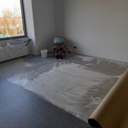 Marek Turowski Usługi Ogólnobudowlane - Pracownik na kolanach rozprowadza klej pod ciemną wykładzinę podłogową w jasnym pomieszczeniu, widoczny zwój wykładziny i wiadro z klejem w rogu pokoju.