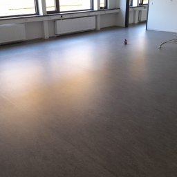 Marek Turowski Usługi Ogólnobudowlane - Nowa, ciemnoszara wykładzina podłogowa w pustym pomieszczeniu biurowym z grzejnikami pod oknami i przewodami wystającymi z podłogi.