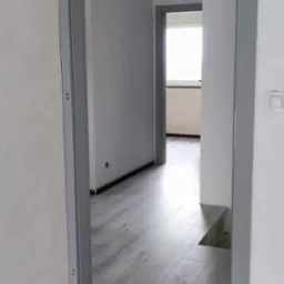 Szare panele podłogowe ułożone w jasnym wnętrzu, widoczne ościeżnice drzwi, surowy stan wykończenia