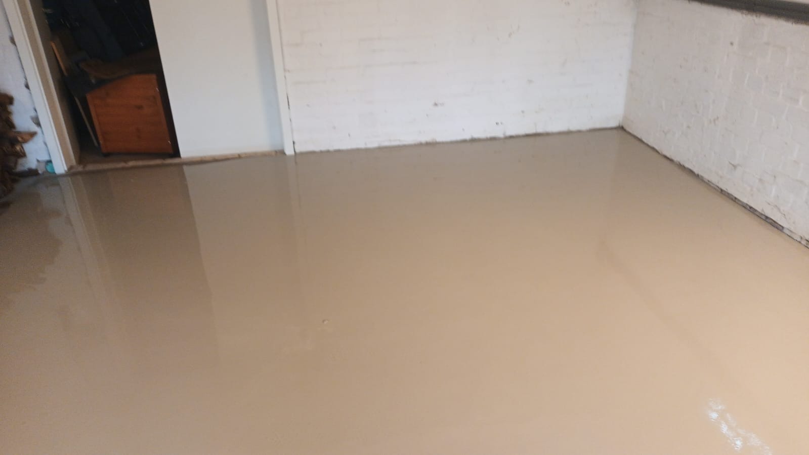 Wylany samopoziomujący podkład podłogowy w pomieszczeniu z białymi ścianami z cegły. Widoczny fragment drewnianej szafki w przejściu do innego pomieszczenia.